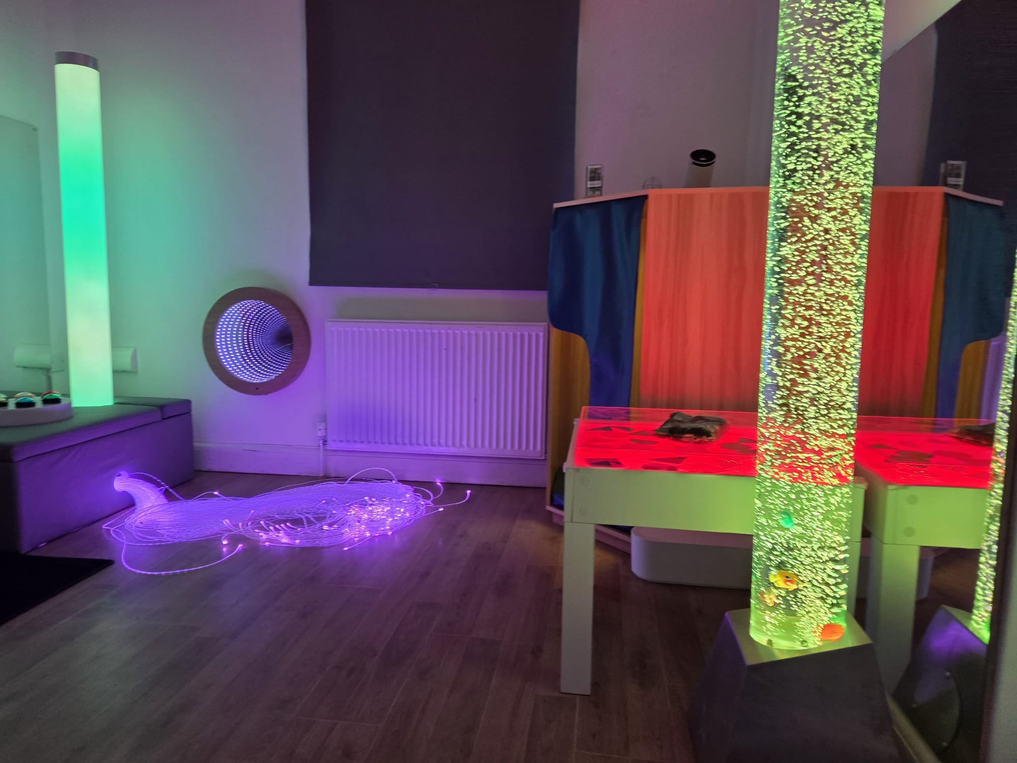 The sensory room lit in green and purple, with a light table, bubble tube and soft seating