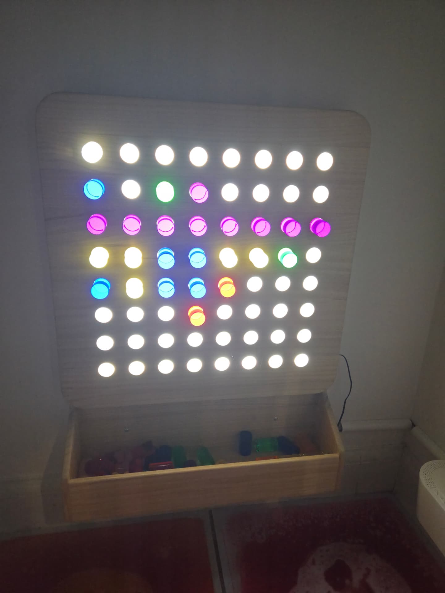 An illuminated wooden peg board with coloured translucent pegs glowing in pink, blue, green and amber